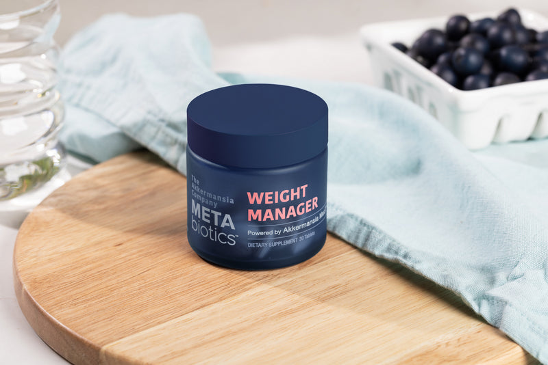 Navy jar of METAbiotics Weight Manager dietary supplement on wooden cutting board with blueberries and teal cloth